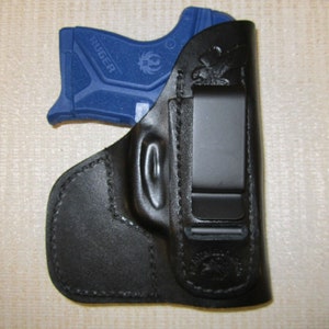 May include: A black leather holster with a clip for carrying a small blue handgun. The gun has the word "RUGER" printed on the side.