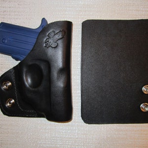 May include: A black leather holster with a blue gun inside. The holster has two silver snaps on the side and a small eagle design on the front. The holster is next to a black leather magazine pouch with two silver snaps.