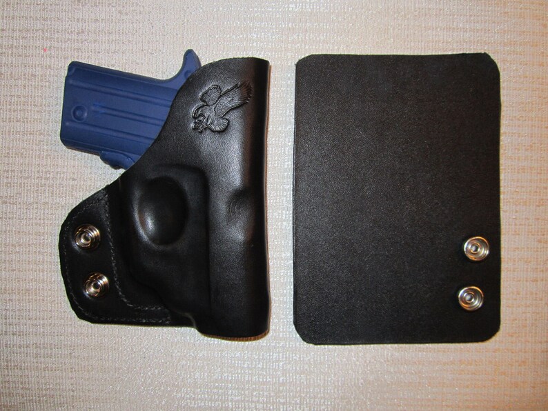 May include: A black leather holster with a blue gun inside. The holster has two silver snaps on the side and a small eagle design on the front. The holster is next to a black leather magazine pouch with two silver snaps.