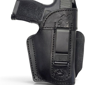 May include: A black leather holster containing a black semi-automatic pistol. The holster features a metal belt clip and an embossed eagle emblem at the top, with a circular logo at the bottom containing the text "BRAZOS HOLSTERS".