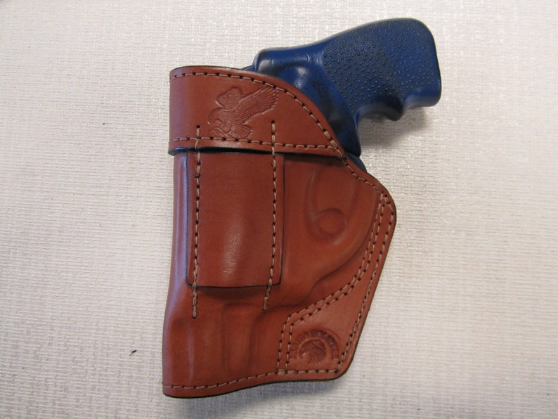 May include: A brown leather holster with a stitched design and an eagle emblem. The holster is designed to hold a blue handgun.