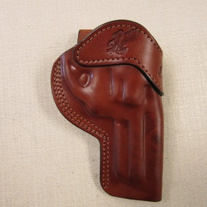 May include: A brown leather holster with a stitched design and an eagle embossed on the top flap.