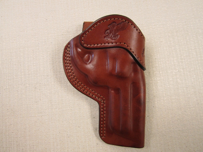 May include: A brown leather holster with a stitched design and an eagle embossed on the top flap.