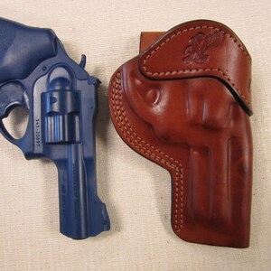 May include: A blue revolver handgun with a leather holster. The holster features a decorative eagle design.