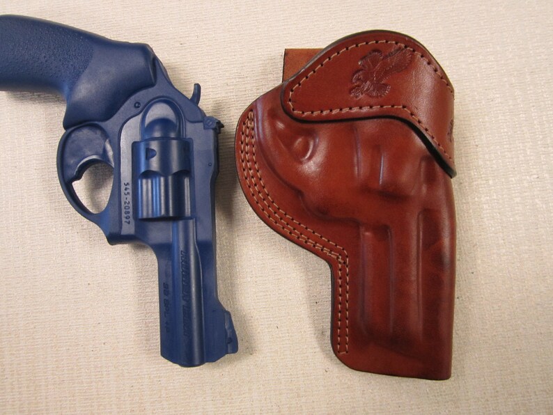 May include: A blue revolver handgun with a leather holster. The holster features a decorative eagle design.