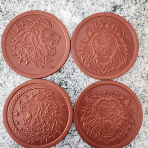 May include: Four round, brown coasters with embossed lion designs. Each coaster features a different lion illustration, including a roaring lion, a lion wearing a crown, and a lion with stars and a crescent moon. The coasters are made of a durable material.