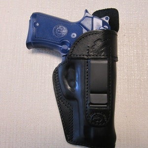 May include: A black leather holster with a belt clip for a handgun. The holster has a stitched design and a logo on the bottom.