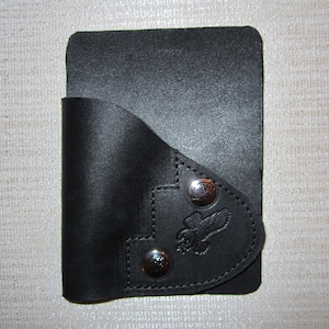 May include: Black leather wallet with a flap and a small pocket on the front. The pocket has a metal snap closure and a decorative eagle design.