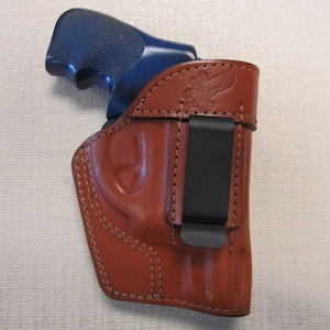 May include: A brown leather holster with a black clip for a handgun. The holster has an eagle design embossed on the side.