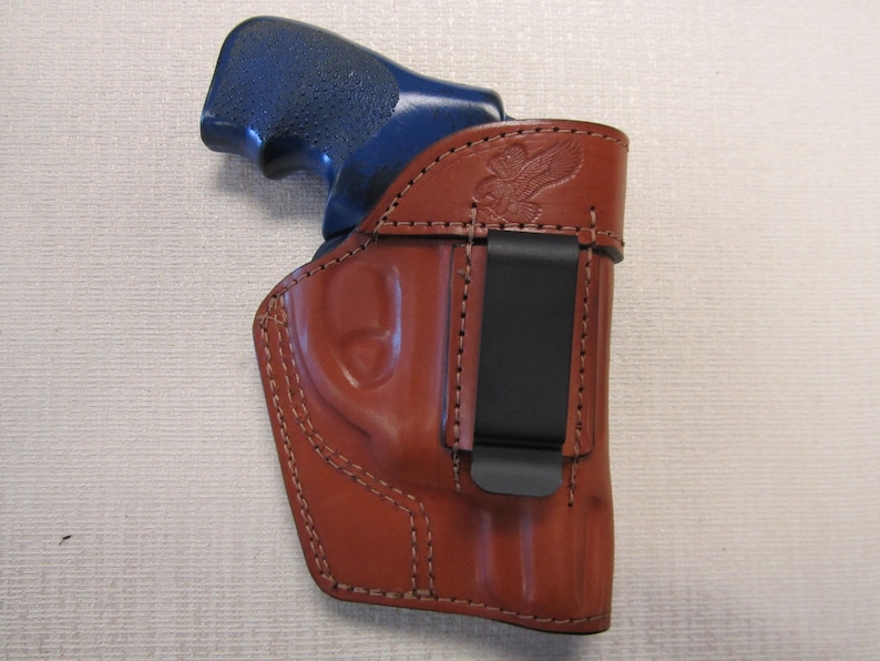May include: A brown leather holster with a black clip for a handgun. The holster has an eagle design embossed on the side.