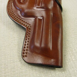 May include: A brown leather holster with stitching around the edges. The holster is designed to hold a handgun.