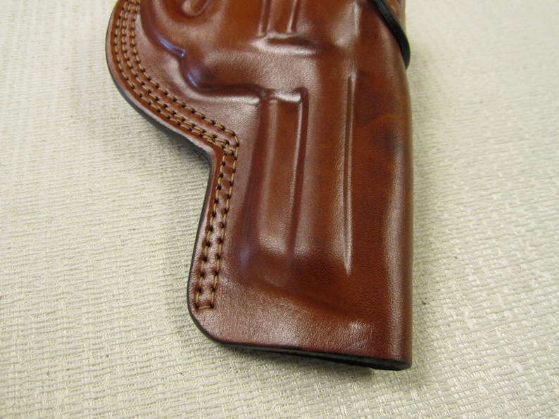 May include: A brown leather holster with stitching around the edges. The holster is designed to hold a handgun.