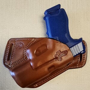 May include: A brown leather holster with a stitched design and an eagle emblem. A blue handgun is partially visible in the holster.