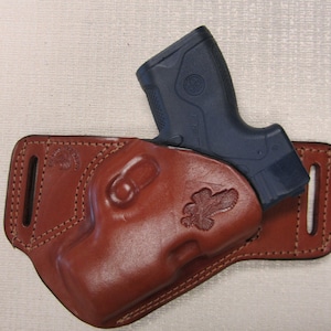 May include: A brown leather holster with a blue handgun inside. The holster has a decorative eagle design.