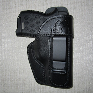 May include: A black leather holster with a clip for a handgun. The holster has a decorative design on the front.