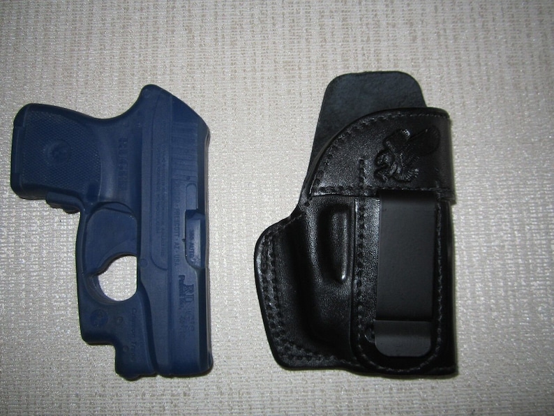 Może przedstawiać: Niebieski pistolet z czarną sk&oacute;rzaną kaburą. Kabura ma klips z tyłu.