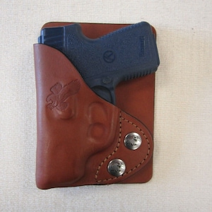 May include: A brown leather holster with a blue handgun inside. The holster has an eagle design and two silver snaps.