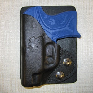 Puede incluir: Una funda de cuero negra con una pistola Ruger azul en su interior. La funda tiene dos broches en forma de &aacute;guila en la parte inferior.