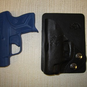 Puede incluir: Una pistola Ruger azul con una funda de cuero negro. La funda tiene dos broches de presi&oacute;n plateados y un dise&ntilde;o cosido.