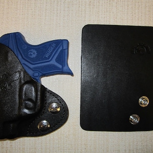 Puede incluir: Una funda de cuero negra con una pistola Ruger azul en su interior. La funda tiene tres broches de presi&oacute;n plateados y un dise&ntilde;o cosido. La funda est&aacute; al lado de un cuadrado de cuero negro con dos broches de presi&oacute;n plateados.