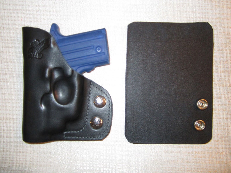 May include: A black leather holster with a blue gun inside. The holster has two silver snaps on the side. There is a black leather flap with two silver snaps on the right side of the image.