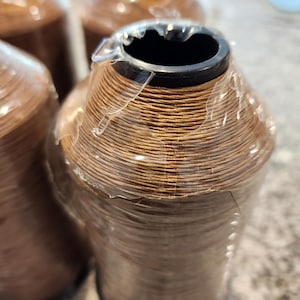 May include: Several spools of brown thread are visible, wrapped in clear plastic. The thread is tightly wound around a black core. The spools are arranged closely together, suggesting a bulk purchase or supply.