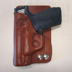 May include: A brown leather holster with a gun inside. The holster has a silver eagle design and two silver snaps.