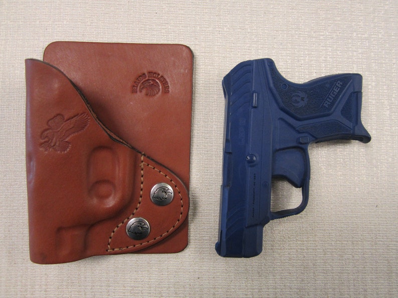 Może przedstawiać: Brązowa sk&oacute;rzana kabura z zatrzaskiem i wytłoczonym logo z przodu. Obok kabury znajduje się niebieski pistolet Ruger.