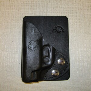 Puede incluir: Funda de cuero negro con dos broches de presi&oacute;n plateados y un logotipo en relieve en la parte delantera.
