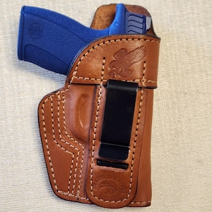 May include: A brown leather holster with white stitching and a black clip. A blue handgun is partially visible inside the holster. The holster features an embossed eagle design and the text "BRAVES HOLSTERS".