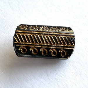 May include: Black glass button with a gold geometric design. The button has a rectangular shape and is decorated with a pattern of lines and swirls.