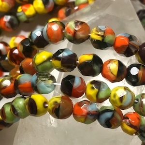 May include: Multicolored glass beads with a faceted finish. The beads are a mix of yellow, orange, red, brown, black, and blue. The beads are strung on a white cord and are displayed on a white crystal.