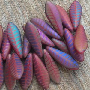 May include: A close-up of a group of red and purple glass beads with a leaf pattern. The beads are iridescent and have a textured surface.