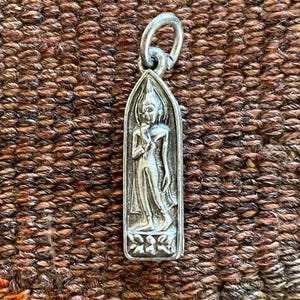 May include: A silver pendant featuring a standing Buddha figure within a rectangular frame. The pendant has a loop at the top for a chain. The background is a textured, brown and orange fabric.