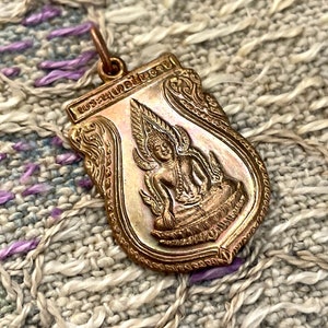 May include: A copper-colored pendant with a detailed image of a seated figure with a halo and ornate details. The pendant has a shield shape with a loop at the top for a chain. The background is a textured fabric.