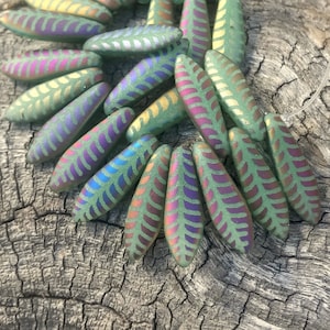 May include: A group of iridescent green glass beads with a leaf pattern. The beads are arranged in a fan shape on a piece of wood.