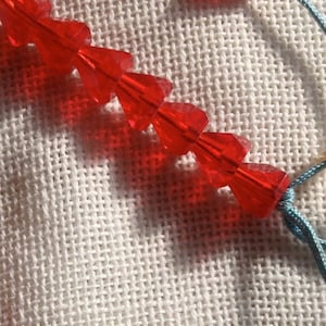 May include: Two strands of red faceted glass beads on a white fabric background. The beads are strung on a blue cord with a white tag attached.