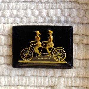 May include: A black and gold enamel pin featuring a couple riding a tandem bicycle. The pin is rectangular and has a black background with gold accents.