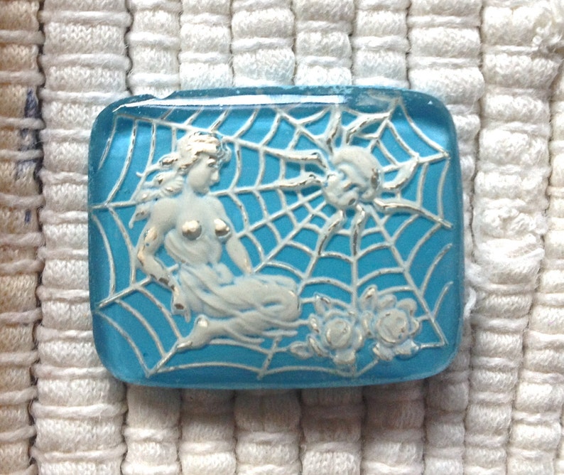 May include: A blue glass tile with a white spider and a woman in a web design.