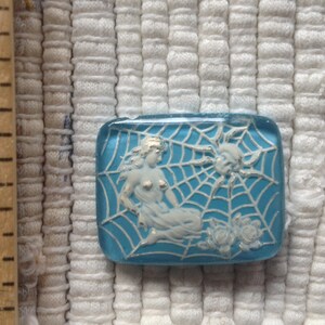 May include: A blue glass button with a white design of a woman in a spider web. The woman is wearing a white dress and has long hair. The spider web is detailed and has a spider in the corner.
