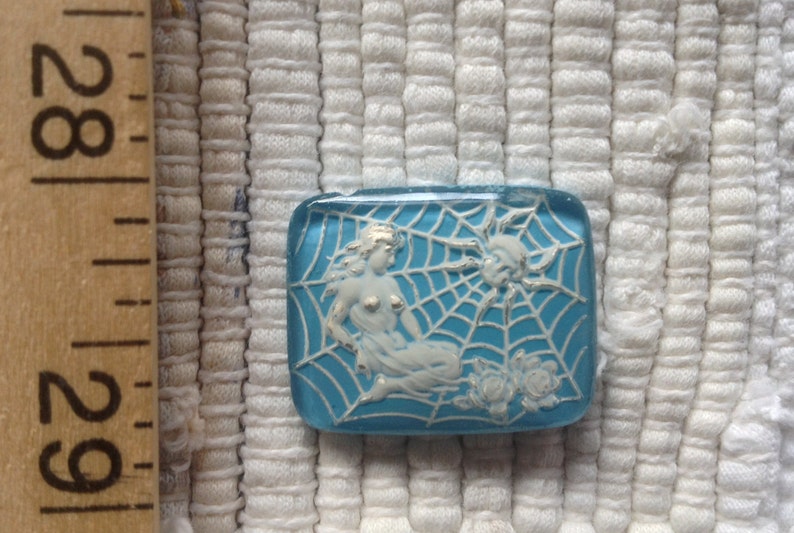 May include: A blue glass button with a white design of a woman in a spider web. The woman is wearing a white dress and has long hair. The spider web is detailed and has a spider in the corner.