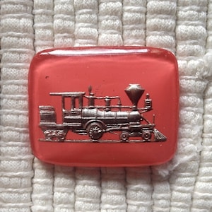 Könnte beinhalten: Ein roter Glasknopf mit einem silberfarbenen Zugmotiv. Der Zug hat einen großen Schornstein und Räder.