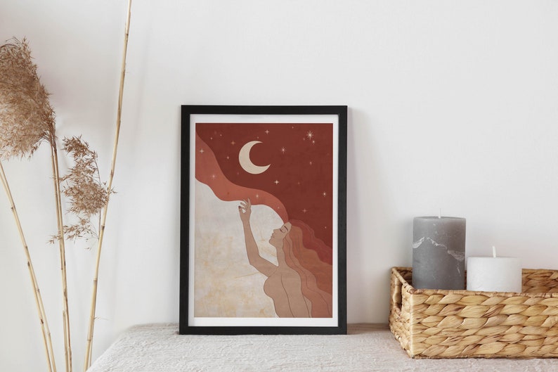 May include: A framed print of a woman with long red hair reaching up towards a crescent moon in a starry night sky. The background is a gradient of red, orange, and white.