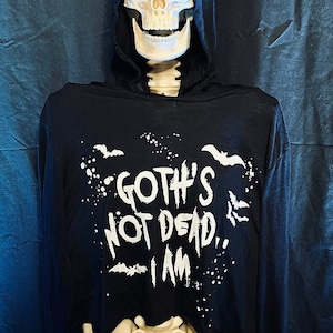 May include: A skeletal figure wearing a black hooded sweatshirt. The sweatshirt features the text "Goth's Not Dead, I Am" in white, along with bat graphics. The figure is posed against a dark blue backdrop.