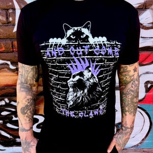 Puede incluir: Camiseta negra con un diseño gráfico que presenta un gato mirando por encima de una pared de ladrillos. El texto "AND OUT GONE" está encima de la pared y "THE CLAWS" debajo. El gato tiene una cresta morada.