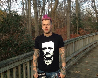 Può includere: T-shirt nero con grafica bianca di Edgar Allan Poe. La persona indossa jeans blu con toppe e abbellimenti. La persona ha tatuaggi sulle braccia e un'acconciatura rosa. La persona è in piedi su un ponte di legno.