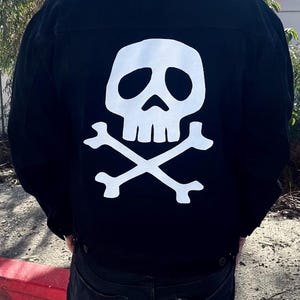 May include: Black denim jacket with a white skull and crossbones graphic on the back. The jacket has a classic button-up design and is worn by a person. The skull and crossbones are a bold, contrasting design element.