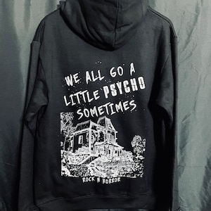 Puede incluir: Sudadera negra con texto blanco que dice "WE ALL GO A LITTLE PSYCHO SOMETIMES". Debajo, un gr&aacute;fico blanco de una casa embrujada y el texto "ROCK N HORROR". La sudadera est&aacute; colgada en una percha de madera.