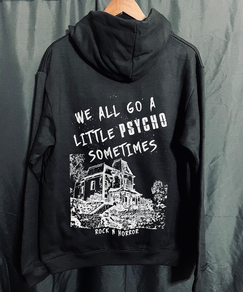 Puede incluir: Sudadera negra con texto blanco que dice "WE ALL GO A LITTLE PSYCHO SOMETIMES". Debajo, un gr&aacute;fico blanco de una casa embrujada y el texto "ROCK N HORROR". La sudadera est&aacute; colgada en una percha de madera.