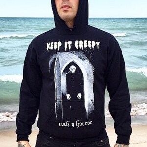May include: Black hooded sweatshirt with the text "KEEP IT CREEPY" and a graphic of a figure in a doorway. The sweatshirt is paired with black jeans featuring skeleton hand patches. The person is wearing sunglasses and standing on a beach.
